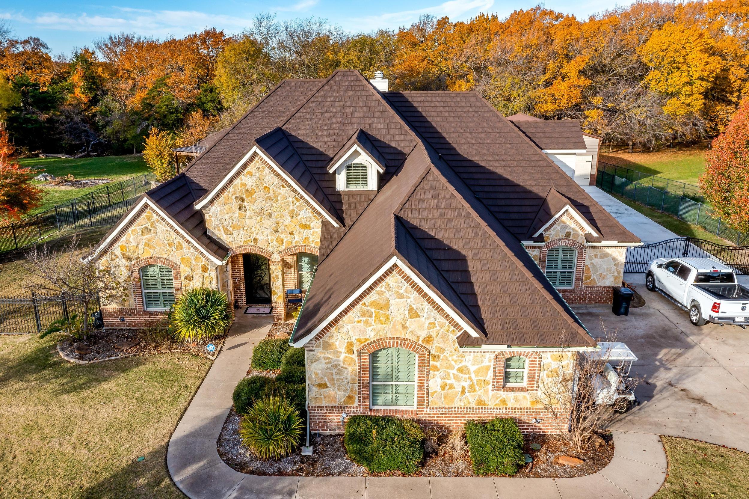 Aerial view of a stone and brick home with a brown stone coated steel roof, autumn trees behind it
